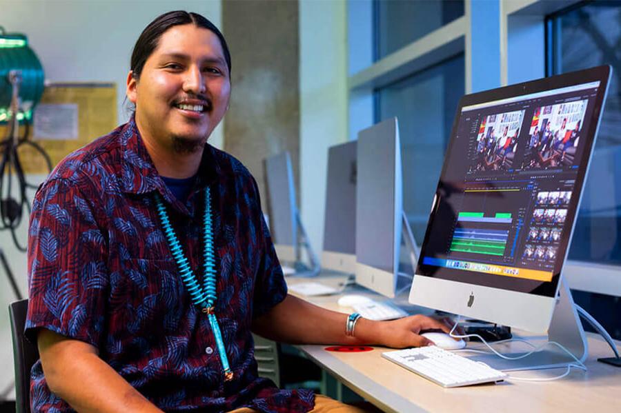 A student sits smiling while wrking on a video project in a student tech lab at Pima West Campus